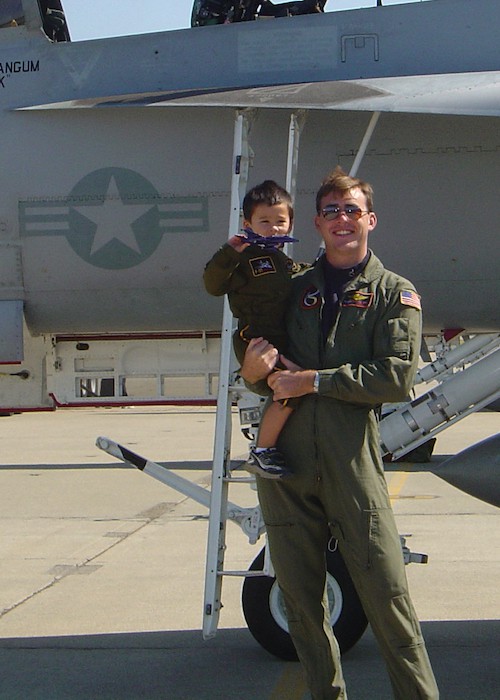 Fighter Pilot Matt Stoll in flight suit holding his young nephew posing in front of a fighter jet
