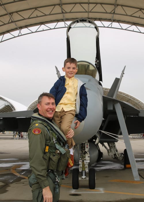 Matt Stoll in his flight suit posing in front of a fighter jet with his young son on his shoulders