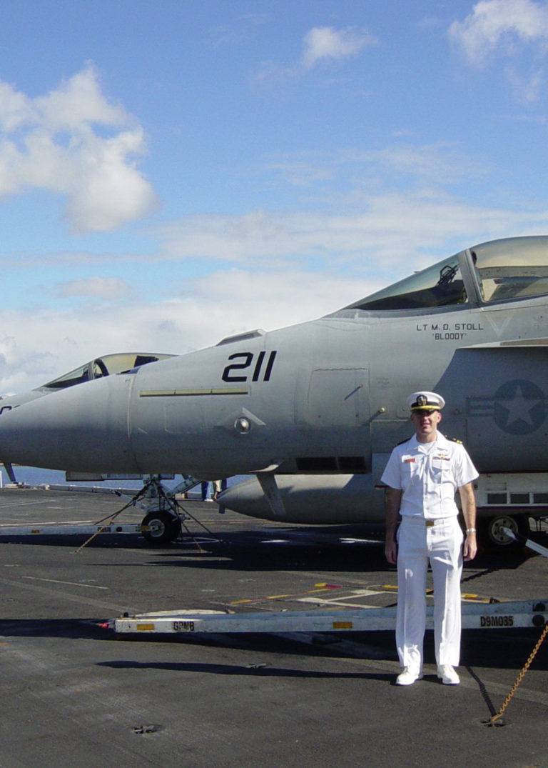 Matt Stoll posing in his dress uniform on an aircraft carrier with jets behind him
