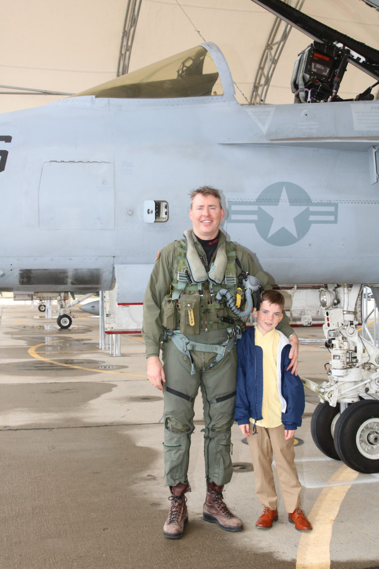Matt Stoll in his flight suit posing with his young son in front of a fighter jet