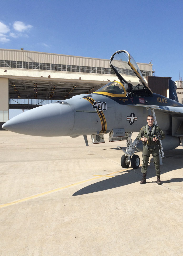 Matt Stoll in a flight suit standing in front of a fighter jet with an airplane hanger behind